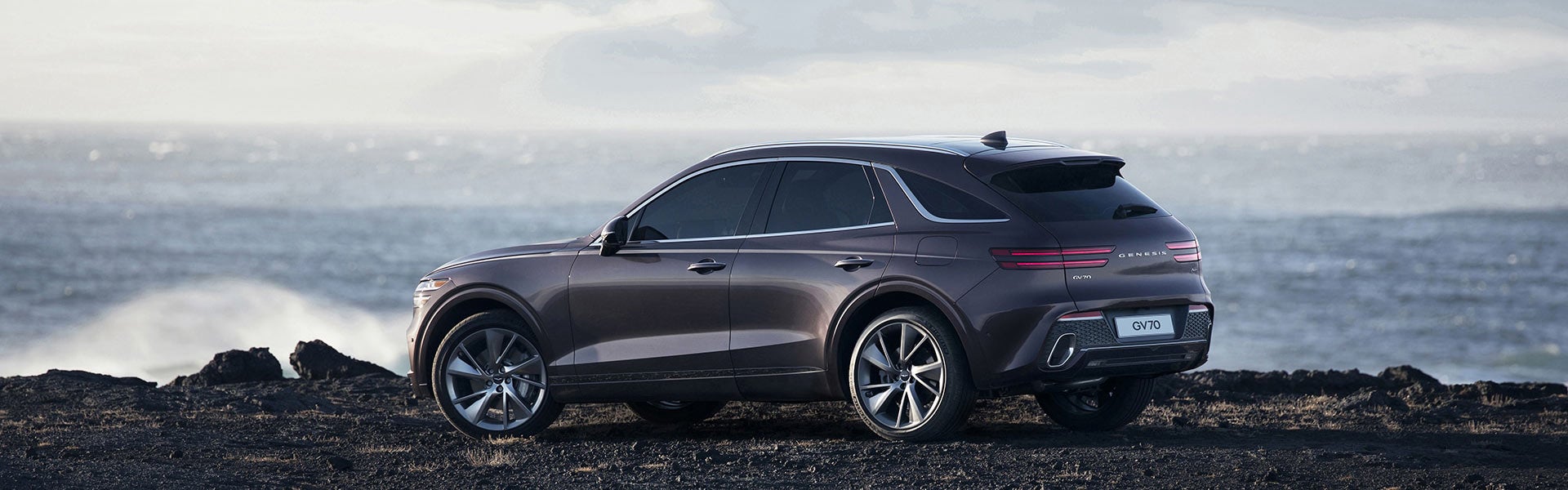 A sleek, Genesis GV70 SUV parked on rocky terrain with ocean waves in the background.