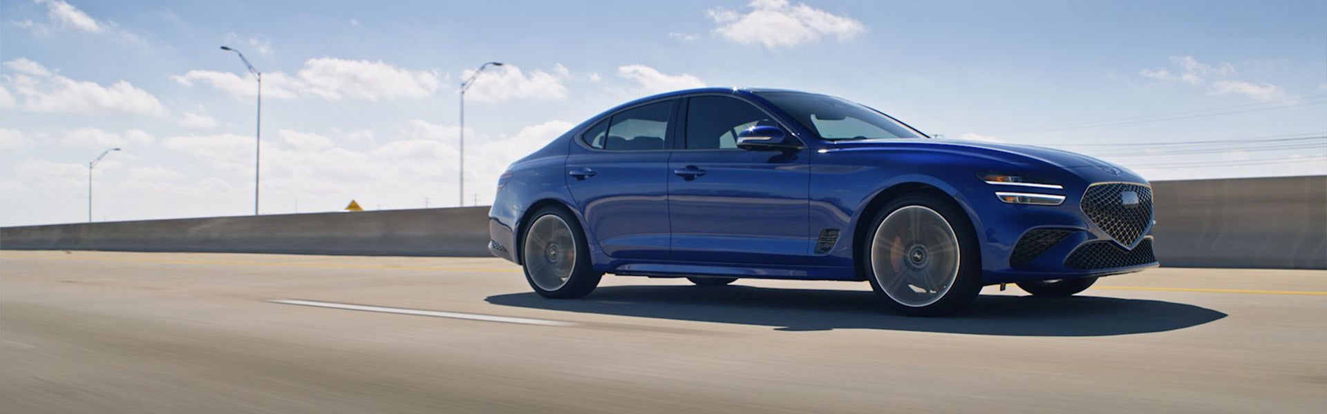 A blue Genesis G70 driving on a highway under a clear sky.