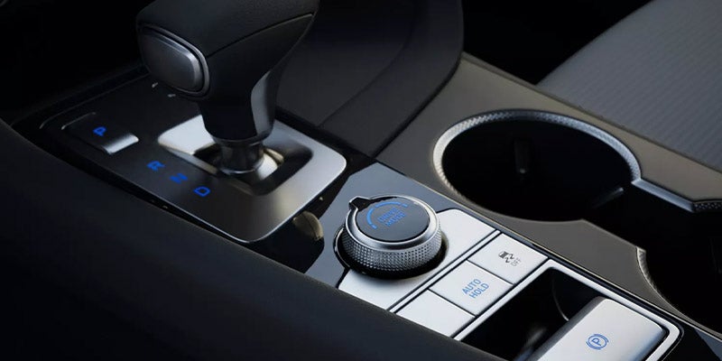 Close-up of a car's center console featuring the gear shifter, control buttons with blue backlighting, and a circular dial.