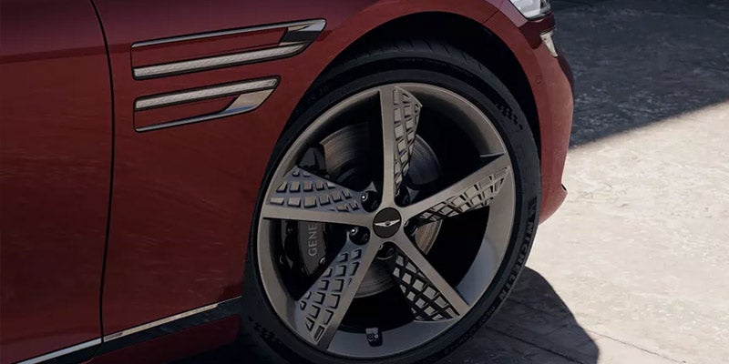 Close-up of a Genesis G80’s wheel featuring a sleek silver alloy rim and a branded brake caliper, set against the backdrop of the vehicle's body.