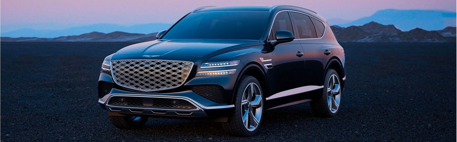 A sleek black 2026 Genesis GV80 with a distinctive grille and modern design parked on a rocky landscape, with mountains in the background during twilight.