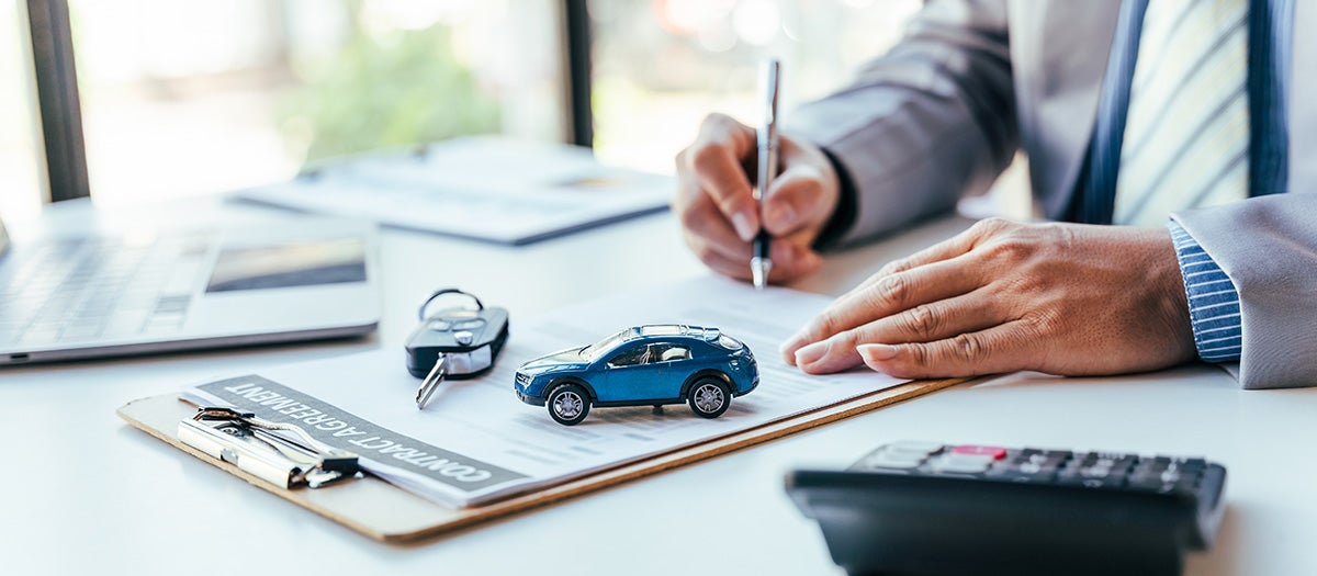 Person signing car insurance documents with car model, keys, and calculator on desk.