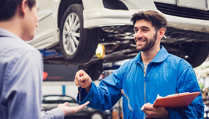 A mechanic in blue coveralls hands over car keys to a customer in front of a vehicle being serviced on a lift.